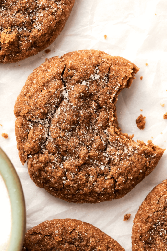 Gros plan d'un biscuit de mélasse de gingembre avec un dessus craquelé enrobé de sucre et une bouchée prise, montrant son intérieur moelleux. D'autres biscuits sont visibles sur du papier sulfurisé en arrière-plan.