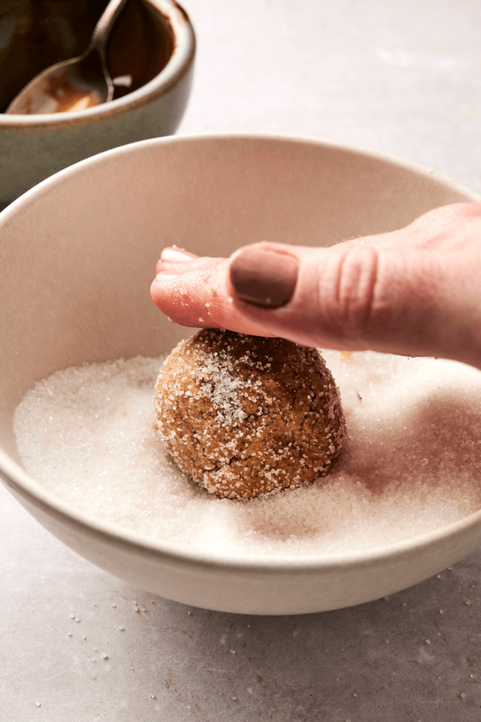 Une main pressant une boule de pâte à biscuits de mélasse de gingembre dans un bol de sucre granulé, la préparant pour la cuisson. Un petit bol avec une cuillère est visible en arrière-plan.