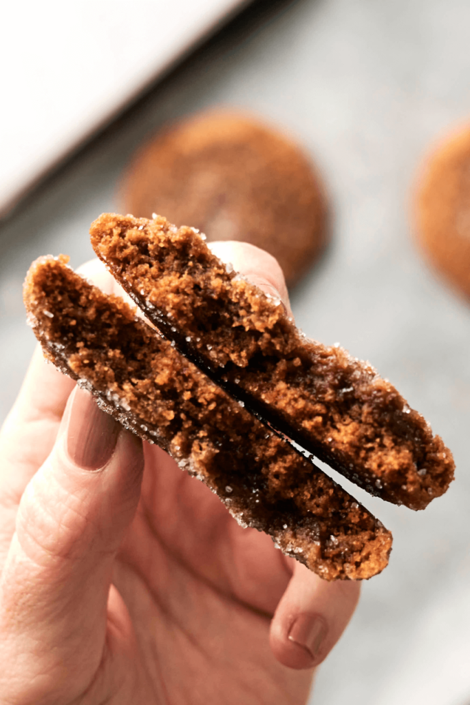 Gros plan d'une main tenant un biscuit de mélasse de gingembre coupé en deux, mettant en valeur son intérieur moelleux et chewy avec une croûte enrobée de sucre. Des biscuits cuits flous sont visibles au fond.