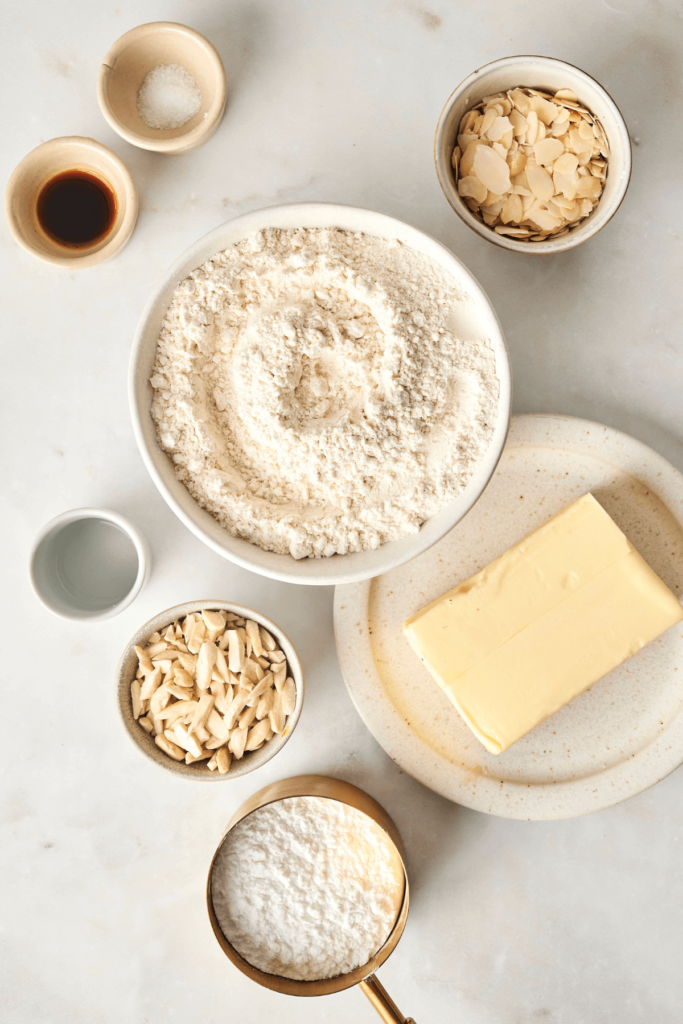 Vue du dessus des ingrédients pour les Biscuits Boule Neige aux Amandes, y compris la farine tout usage, le sucre glace, les amandes tranchées et émondées, le beurre non salé, l'extrait de vanille, l'extrait d'amande et le sel, disposés sur une surface en marbre blanc.