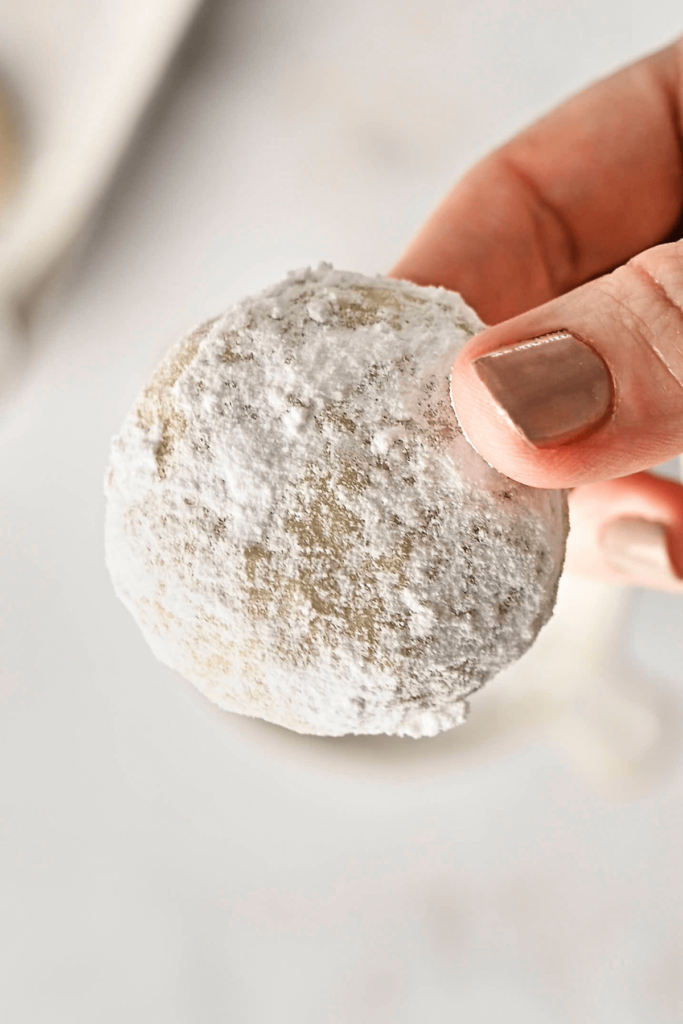 Une main tenant un Biscuit Boule Neige aux Amandes fraîchement enrobé de sucre glace, mettant en valeur son extérieur délicat recouvert de sucre.