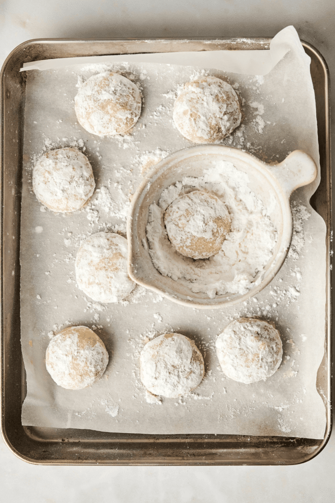 Biscuits Boule Neige tout juste cuits en cours d’enrobage de sucre glace, avec un biscuit reposant dans un bol de sucre glace et d'autres sur une plaque de cuisson tapissée de papier sulfurisé.