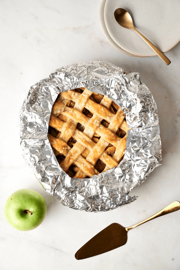 Une tarte aux pommes dorée avec une croûte en treillis entourée d'une protection en aluminium pour éviter un brunissement excessif, présentée avec une pomme verte, un serveur à tarte et des assiettes à proximité.