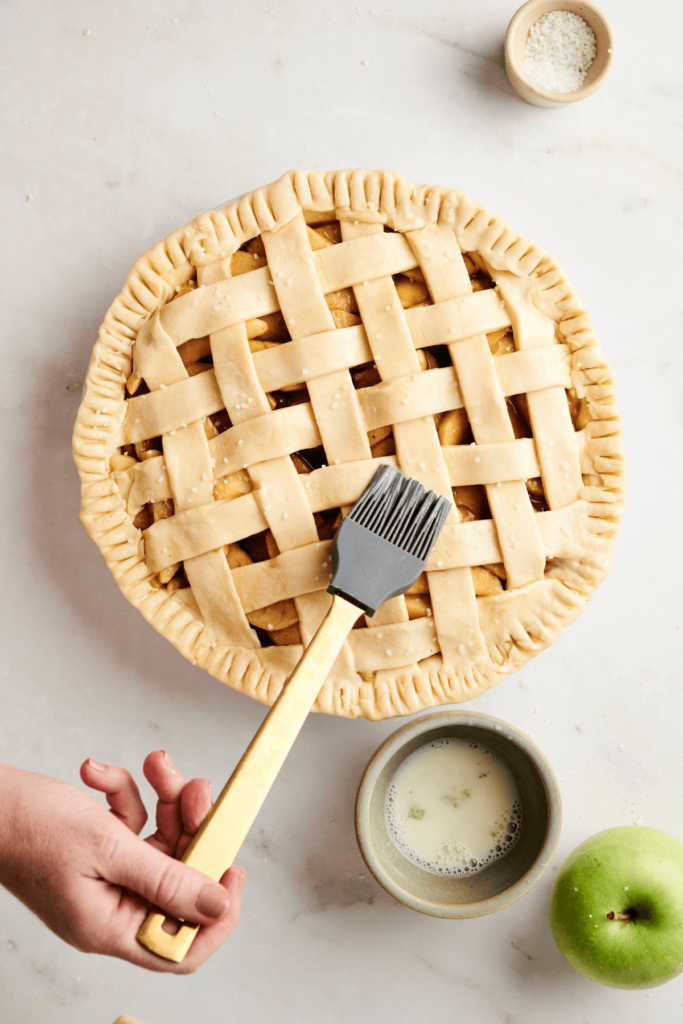 Une tarte aux pommes avec treillis étant badigeonnée de dorure avec un pinceau à poignée dorée, avec un bol de dorure et une pomme verte à proximité.