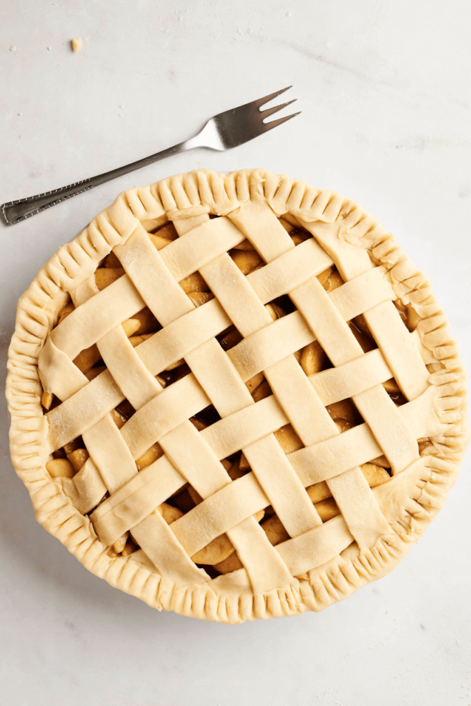 Une tarte aux pommes non cuite avec une croûte en treillis, bords repliés, et une pomme verte fraîche à proximité.