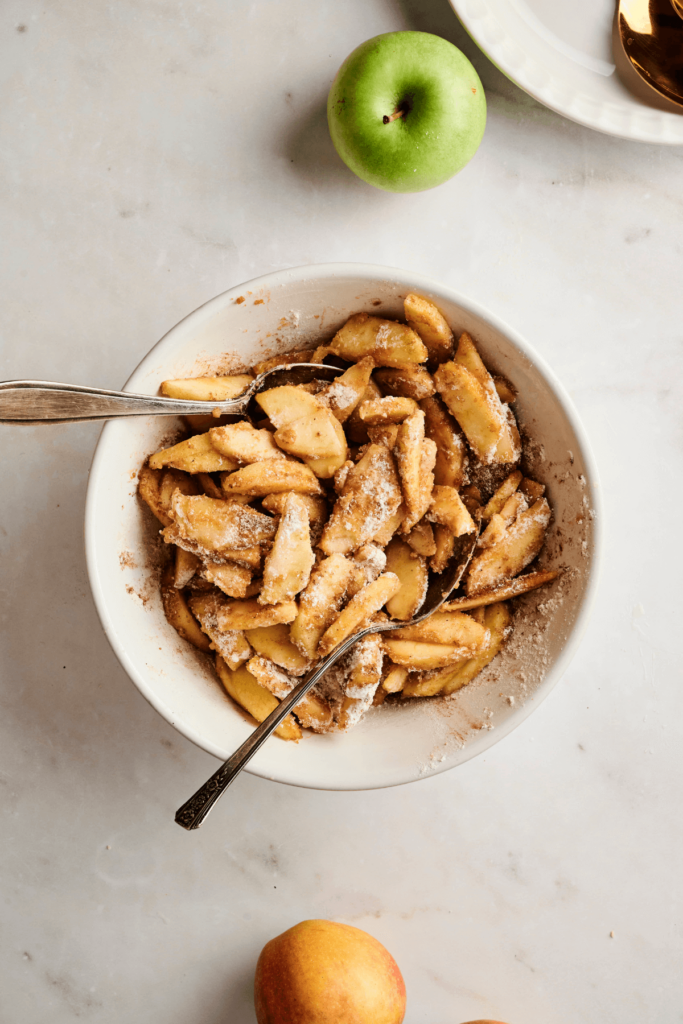 Tranches de pommes fraîches dans un bol enrobées d'un mélange de cannelle, sucre brun et farine, prêtes à être mélangées pour la garniture de tarte aux pommes.