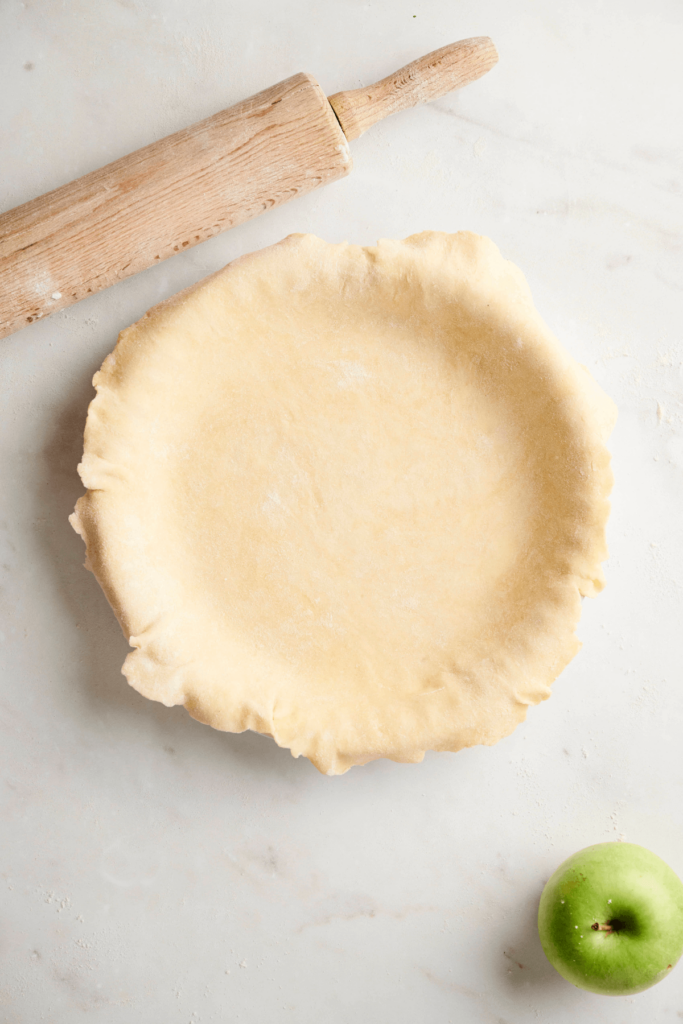 Pâte à tarte adaptée dans un plat à tarte sur une surface blanche.