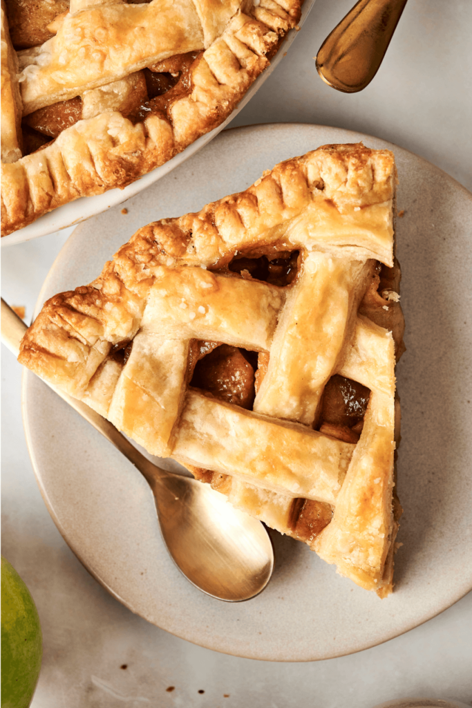 Close-up of a slice of apple pie with a golden, flaky lattice crust, served on a plate with a gold spoon.