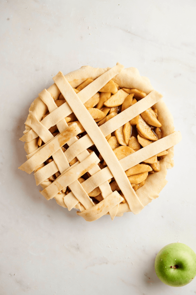 Partially completed lattice pie crust being woven over an apple pie filling, with a green apple placed nearby on a light surface.