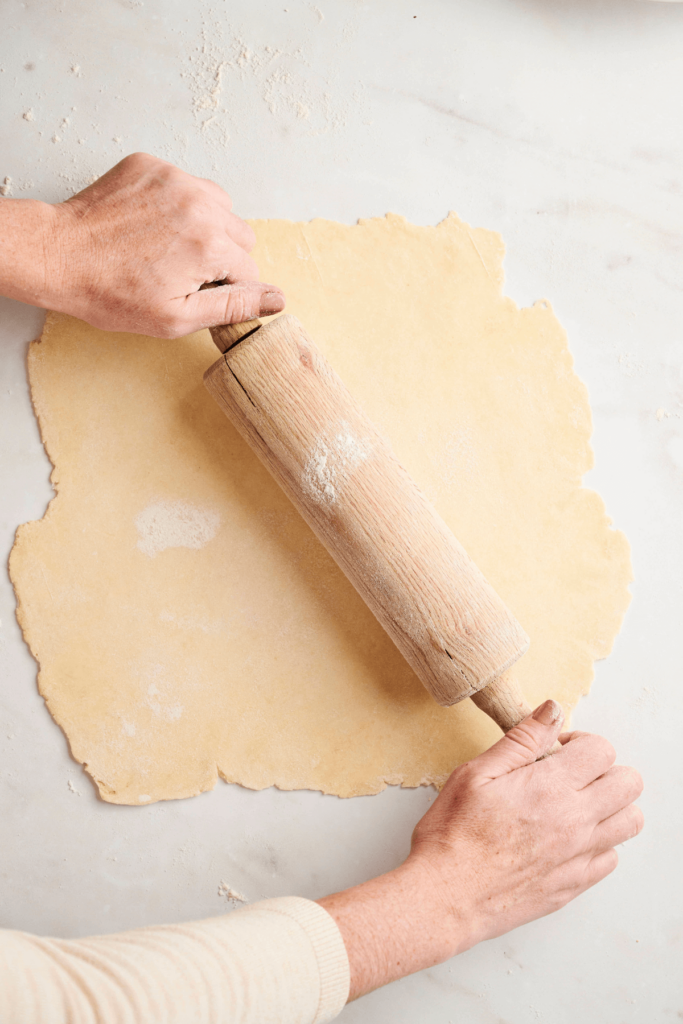 Hands using a rolling pin to evenly roll out all butter pie dough on a floured surface.