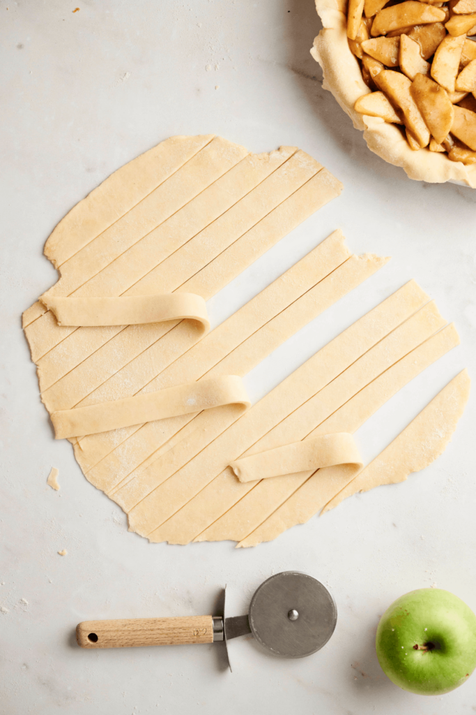 Rolled-out pie dough cut into strips for lattice topping, with a pizza cutter and a green apple nearby.