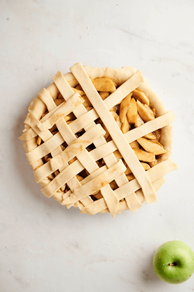 Partially completed lattice pie crust being woven over an apple pie filling, with a green apple placed nearby on a light surface.