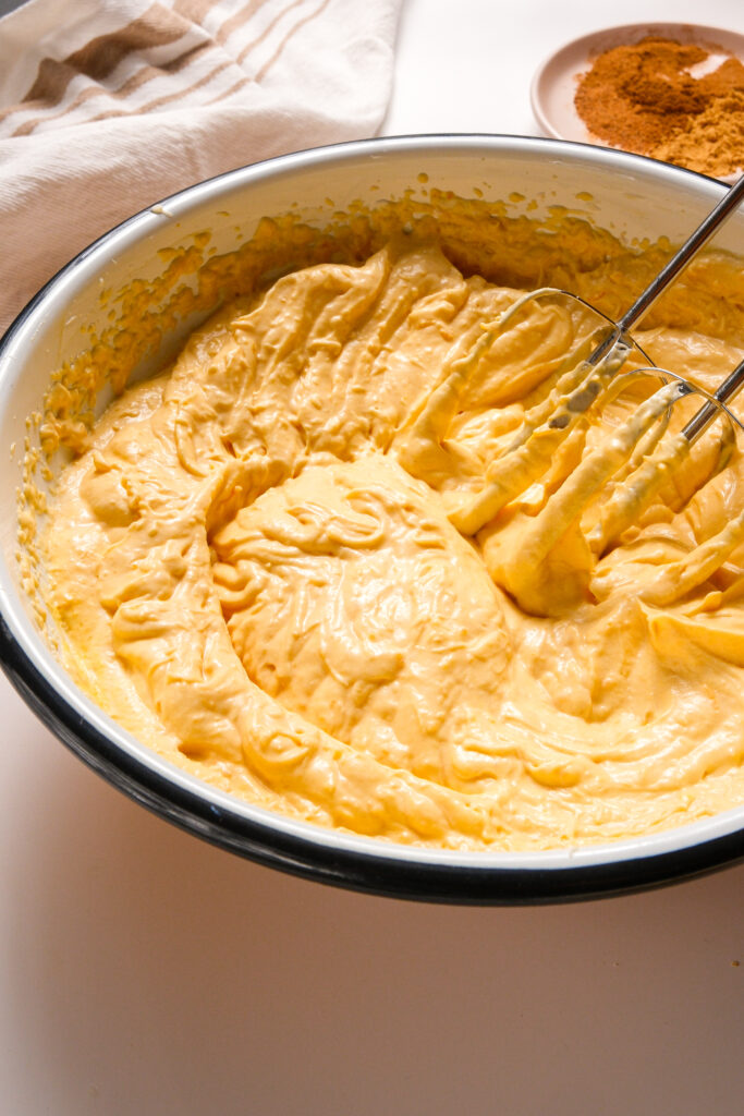 Bol de garniture de cheesecake à la citrouille en cours de mélange avec un batteur à main, comprenant de la purée de citrouille, de la crème aigre, de l'extrait de vanille et du jus de citron.