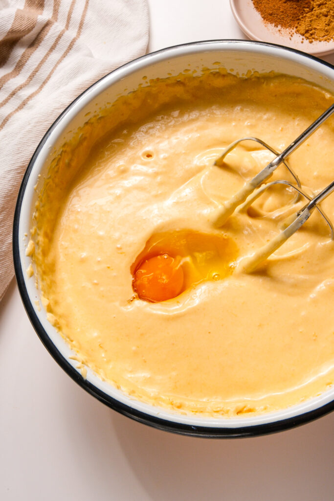 Un œuf ajouté à la pâte de cheesecake à la citrouille dans un bol de mélange, avec un batteur à main prêt à incorporer.
