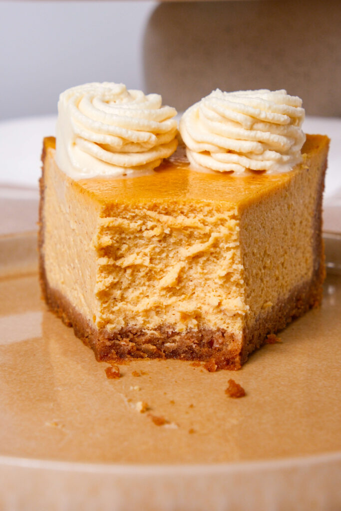 Gros plan sur une part de cheesecake à la citrouille avec une bouchée prise, surmonté de tourbillons de crème fouettée à la cannelle, mettant en valeur la texture crémeuse et la riche croûte en biscuit graham.