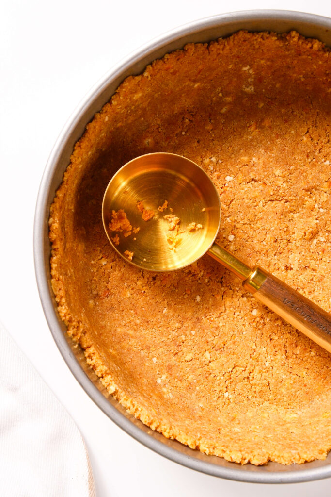 Un moule à charnière avec une croûte de biscuit graham pressée uniformément à l'aide d'une tasse à mesurer pour un cheesecake à la citrouille.