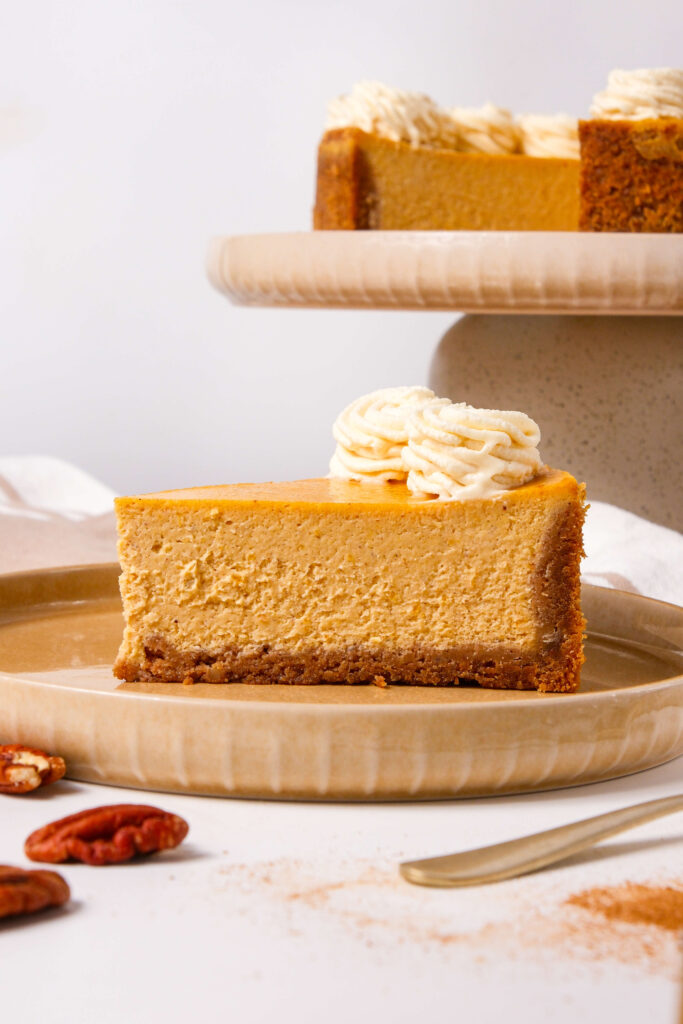 Vue latérale d'une part de cheesecake à la citrouille sur une assiette, avec une croûte de biscuit graham et surmontée de tourbillons de crème fouettée à la cannelle, avec le cheesecake entier visible en arrière-plan.