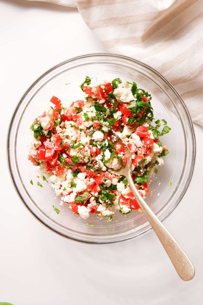 Un bol en verre rempli d'un mélange de tomates hachées, de feta émiettée et de basilic frais, avec une cuillère prête à être utilisée.