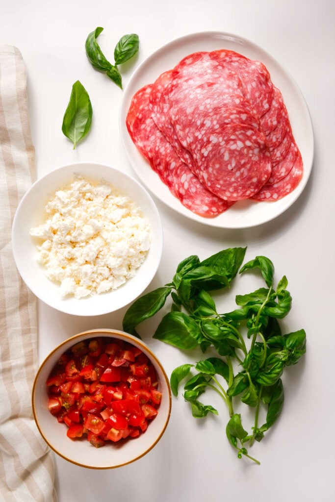 Ingrédients pour les bouchées de salami croustillantes disposés sur une surface blanche, comprenant des tranches de salami sur une assiette, de la feta émiettée dans un bol, des tomates hachées dans un bol et des feuilles de basilic frais.