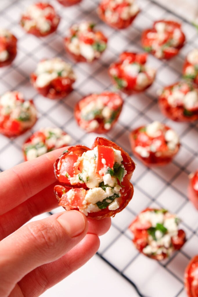 Gros plan sur une main tenant une bouchée de salami croustillante remplie de feta, tomate et basilic, avec d'autres bouchées de salami sur une grille de refroidissement en arrière-plan.