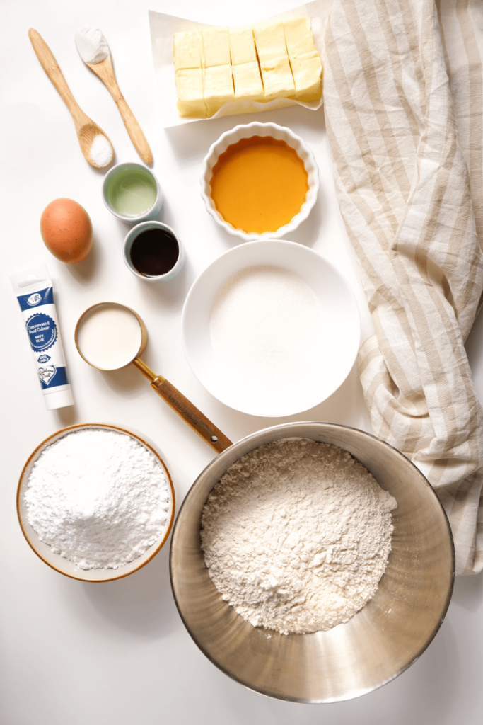 Ingrédients pour les biscuits de Noël au sucre, y compris la farine, le beurre, le sucre, l'œuf, l'extrait d'amande, la poudre à lever et le sel, disposés sur une surface blanche.