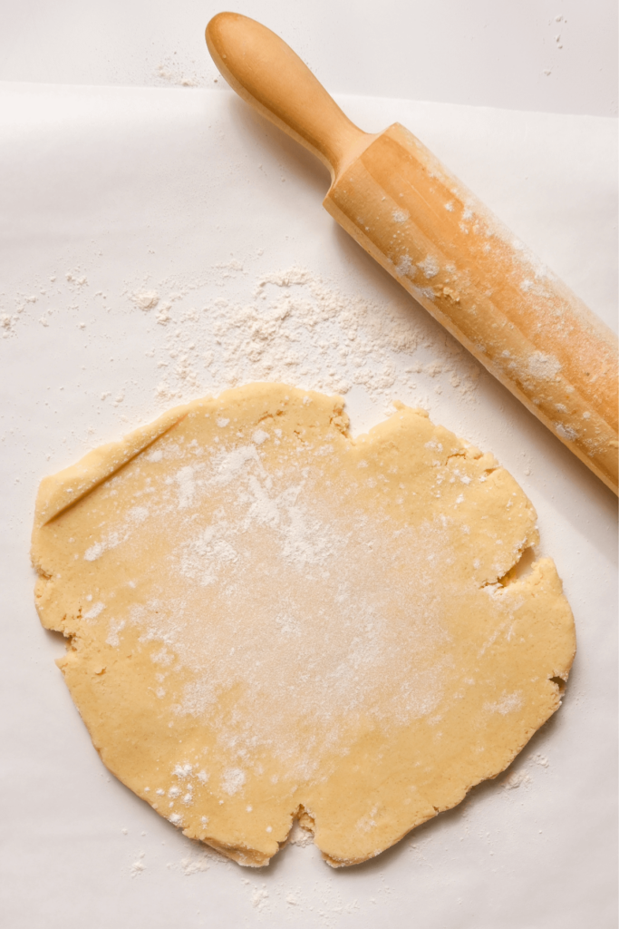 Pâte à biscuits de Noël étalée sur du papier sulfurisé, saupoudrée de farine, accompagnée d'un rouleau à pâtisserie, prête à être découpée en formes.