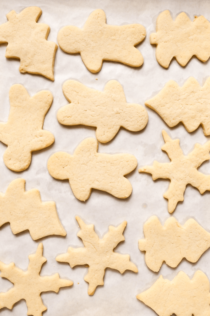 Biscuits de Noël au sucre fraîchement cuits dans diverses formes festives, y compris des arbres, des personnages en pain d'épice et des flocons de neige, sur un plateau de cuisson tapissé de papier sulfurisé.