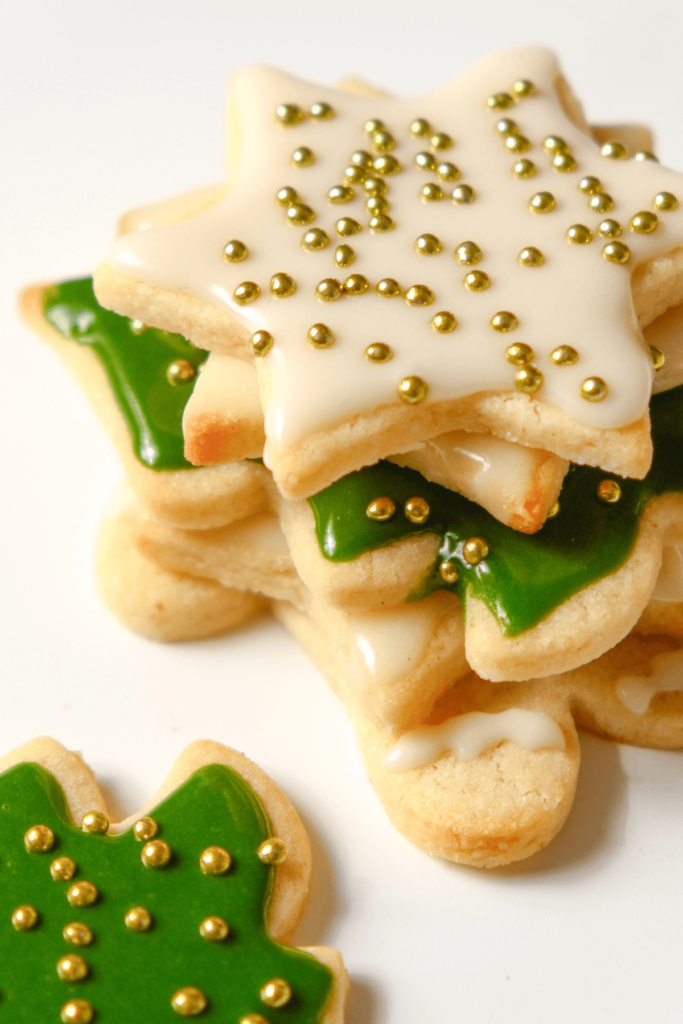 Gros plan d'une pile de biscuits au sucre en forme d'étoile et d'arbre décorés de glaçage blanc et vert, surmontés de vermicelles dorés.