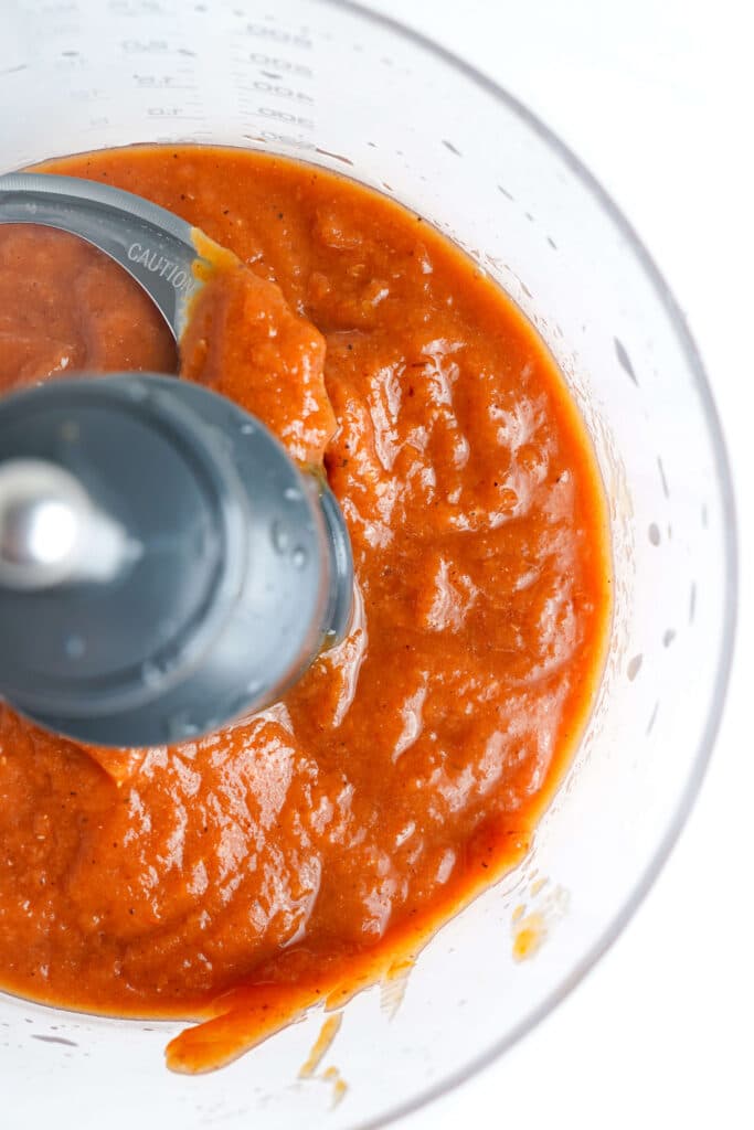 Purée de tomates rôties au feu dans un robot culinaire avec des assaisonnements visibles et une lame partiellement submergée, prête à être utilisée dans une recette.