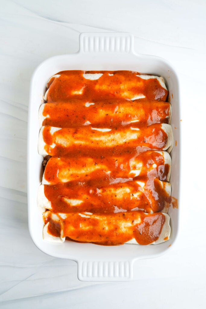 Un plat de cuisson blanc rempli d'enchiladas de bœuf roulées, garnies de sauce enchilada maison, prêtes à être cuites.