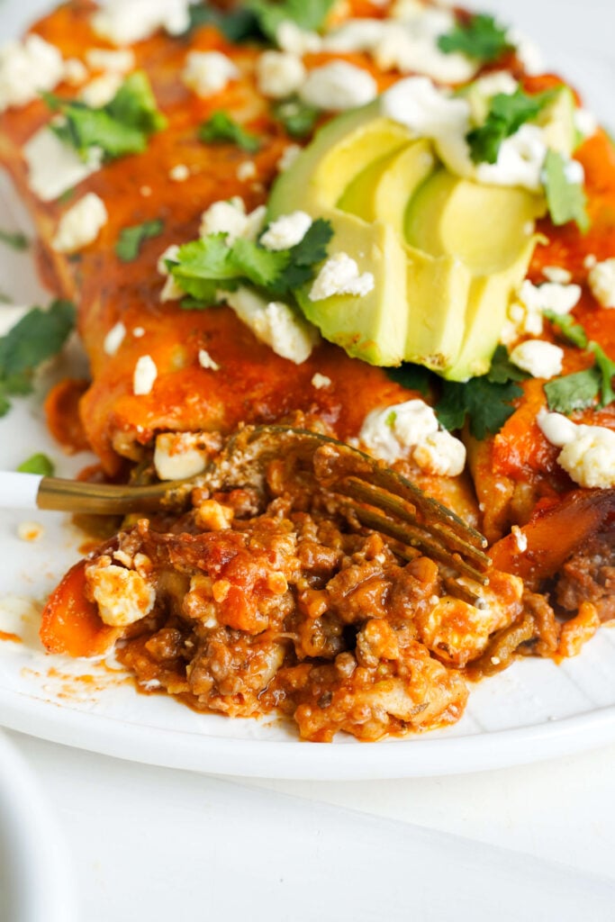 Gros plan d'une enchilada au bœuf au fromage sur une assiette, garnie de tranches d'avocat, de coriandre, et de fromage émietté, avec une fourchette coupant dans la garniture.