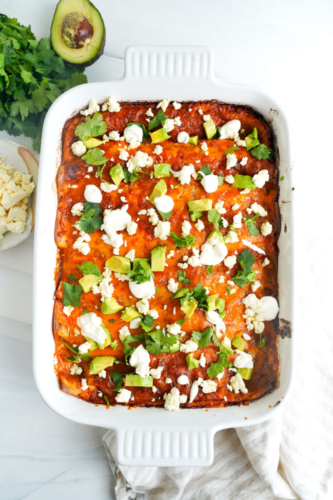 Un plat de cuisson blanc rempli d'enchiladas au bœuf au fromage, garnies d'avocat, de coriandre, de fromage émietté, et de cuillerées de crème aigre.