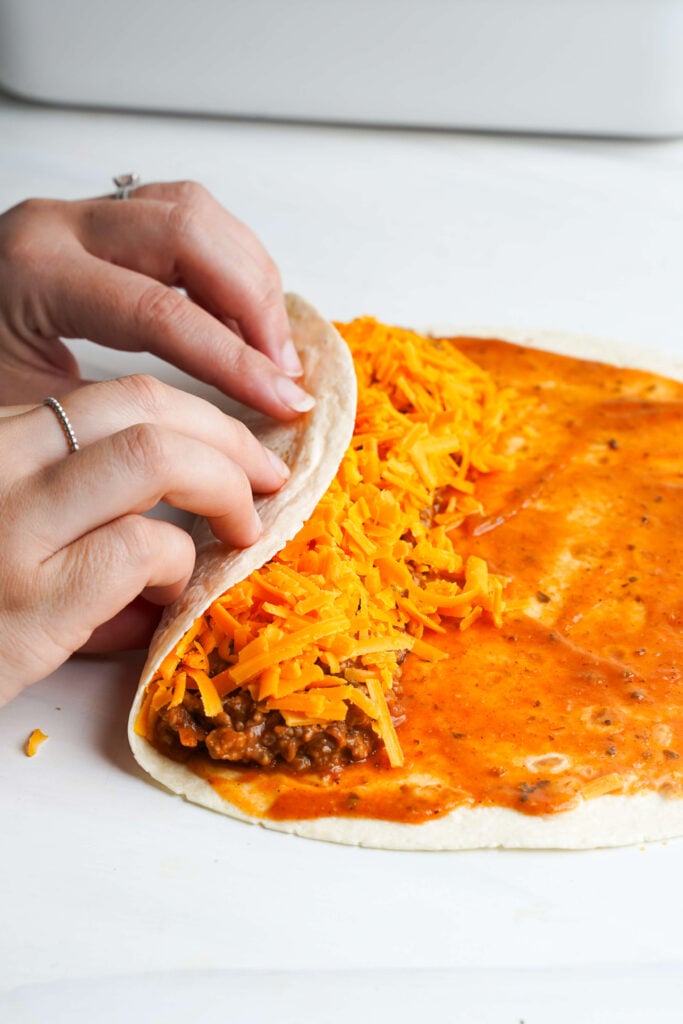 Mains enroulant une tortilla de farine remplie de bœuf assaisonné, fromage cheddar râpé et sauce enchilada sur une surface blanche.