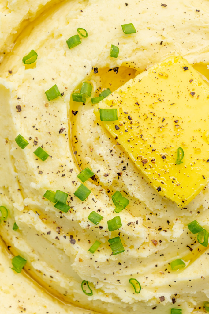 Gros plan sur de la purée crémeuse de pommes de terre surmontée d'un morceau de beurre, de poivre noir fraîchement moulu et d'oignons verts hachés.