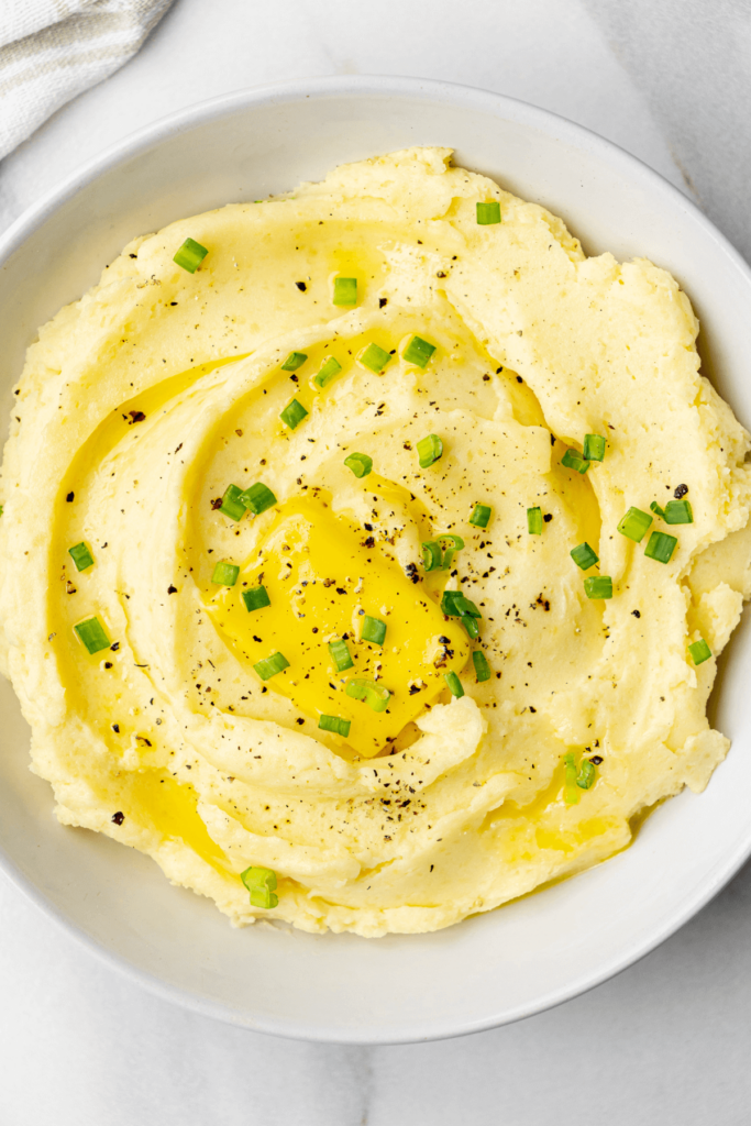 Gros plan sur de la purée de pommes de terre surmontée d'un morceau de beurre, de poivre noir fraîchement moulu et d'oignons verts hachés dans un bol blanc.