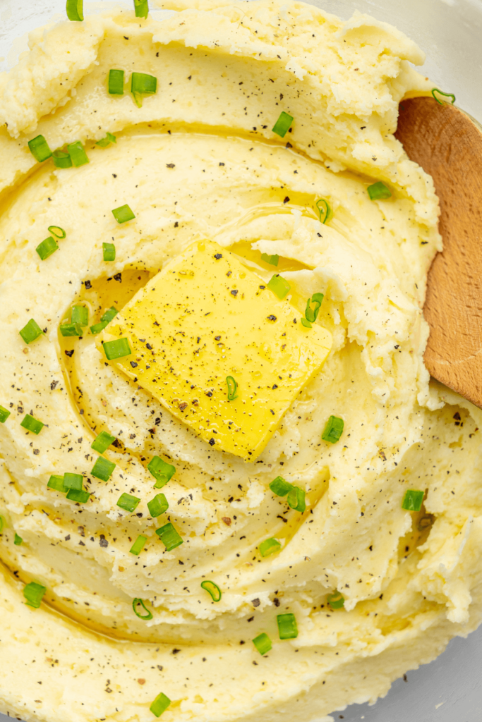 Gros plan sur de la purée de pommes de terre surmontée d'un morceau de beurre, de poivre noir fraîchement moulu et d'oignons verts hachés, prête à être servie.