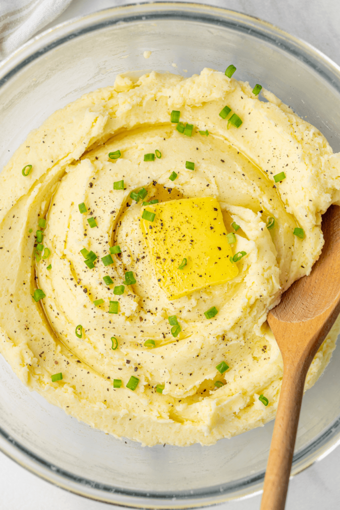 Gros plan sur de la purée crémeuse de pommes de terre surmontée d'un morceau de beurre, de poivre noir fraîchement moulu et d'oignons verts hachés.