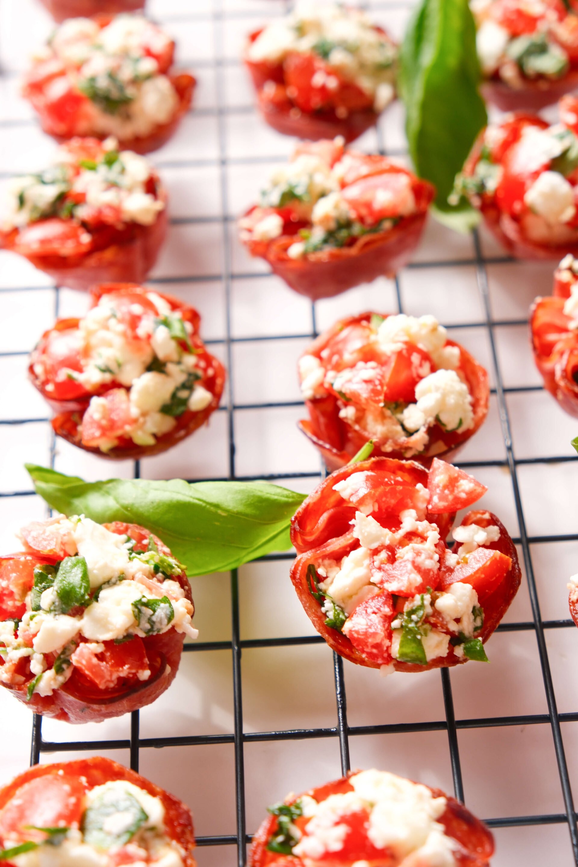 Bouchées de salami croustillantes remplies de feta, tomate et basilic disposées sur une grille de refroidissement, garnies de feuilles de basilic frais.