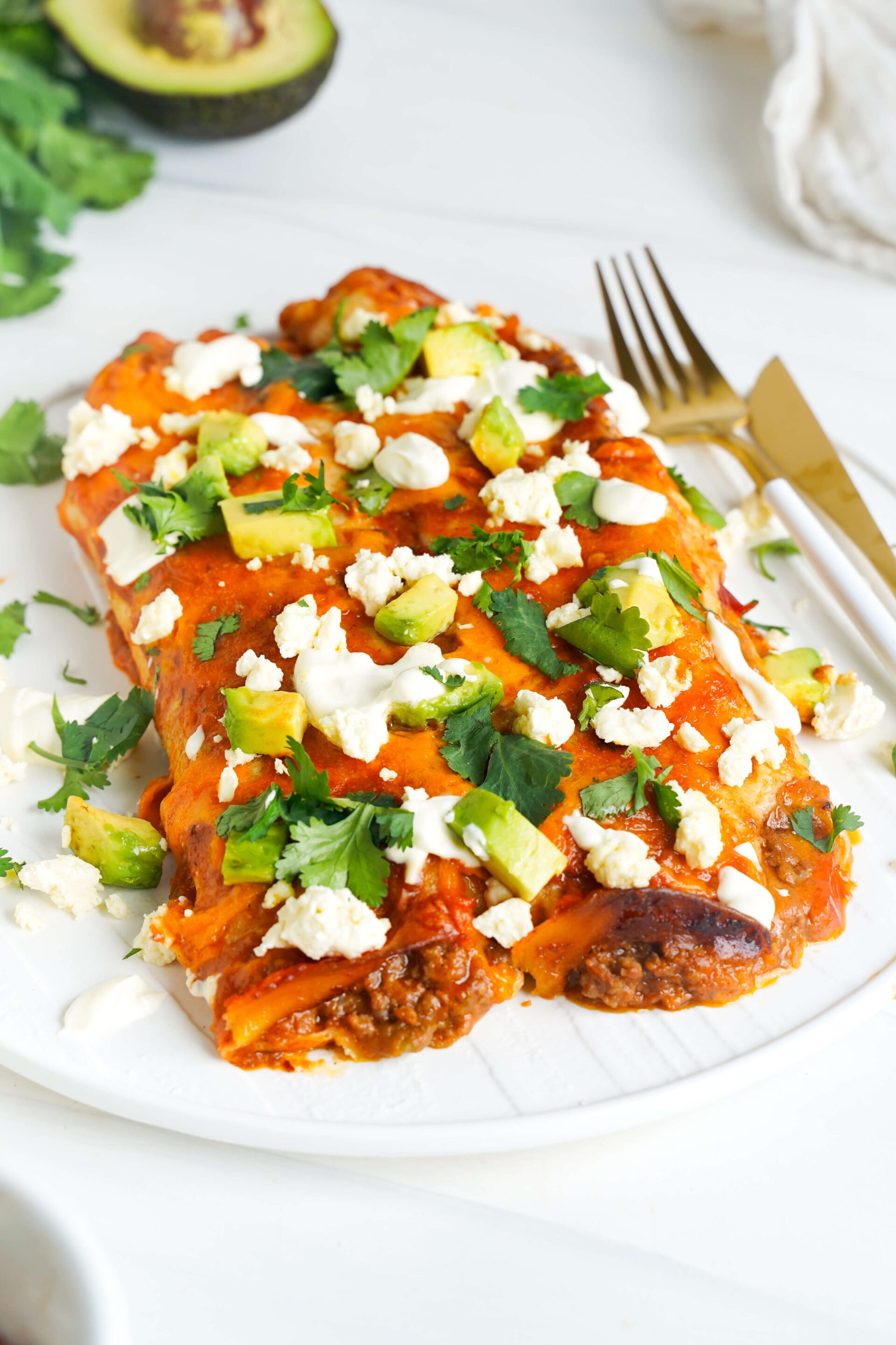 Enchiladas au fromage au bœuf présentées avec de l'avocat, de la coriandre, du fromage émietté et de la crème aigre, servies avec des ustensiles dorés sur une assiette blanche.