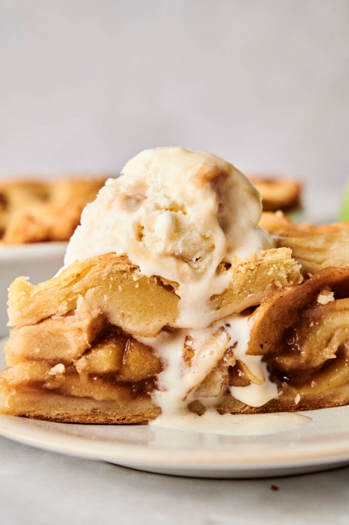 Gros plan d'une part de tarte aux pommes maison avec des couches de pommes tendres épicées à la cannelle et une croûte feuilletée, surmontée de glace à la vanille fondue sur une assiette blanche.