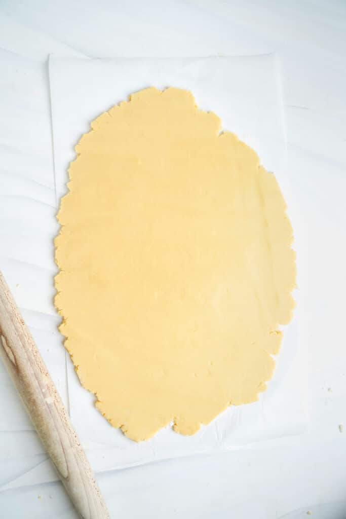 Pâte à biscuit au sucre étalée sur un morceau de papier sulfurisé. Rouleau à pâtisserie à côté.
