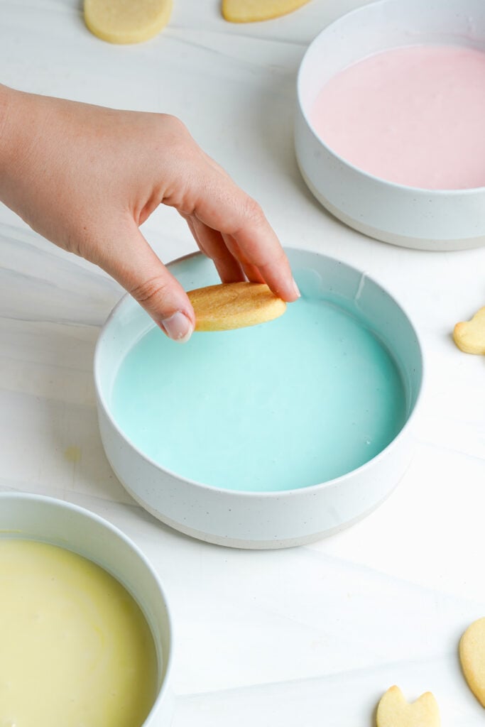 Main trempant un biscuit au sucre de Pâques dans un bol de glaçage bleu pastel pour la décoration.