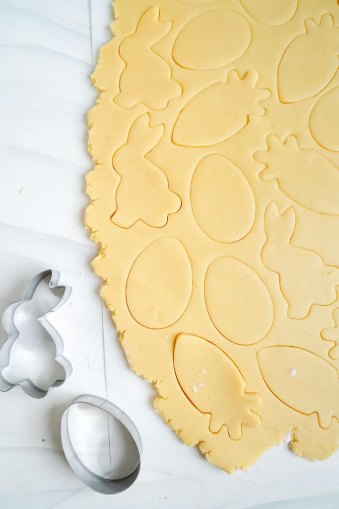 Pâte à biscuit au sucre de Pâques étalée et découpée en formes de lapins, d'œufs et de carottes avec des emporte-pièces en métal à côté.