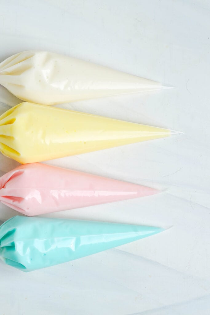Quatre poches de pâtisserie remplies de glaçage pastel de Pâques en blanc, jaune, rose et bleu, prêtes à décorer des biscuits.