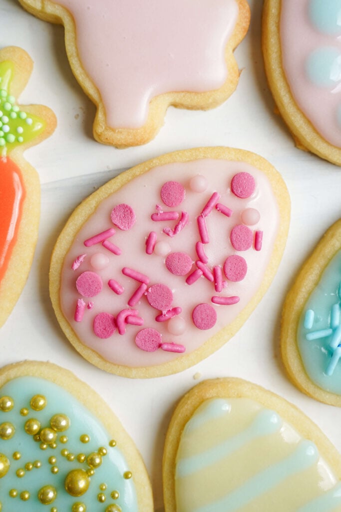 Gros plan d'un biscuit en forme d'œuf de Pâques rose décoré avec un glaçage pastel rose et des vermicelles roses vifs, entouré d'autres biscuits décorés colorés en diverses formes de Pâques.