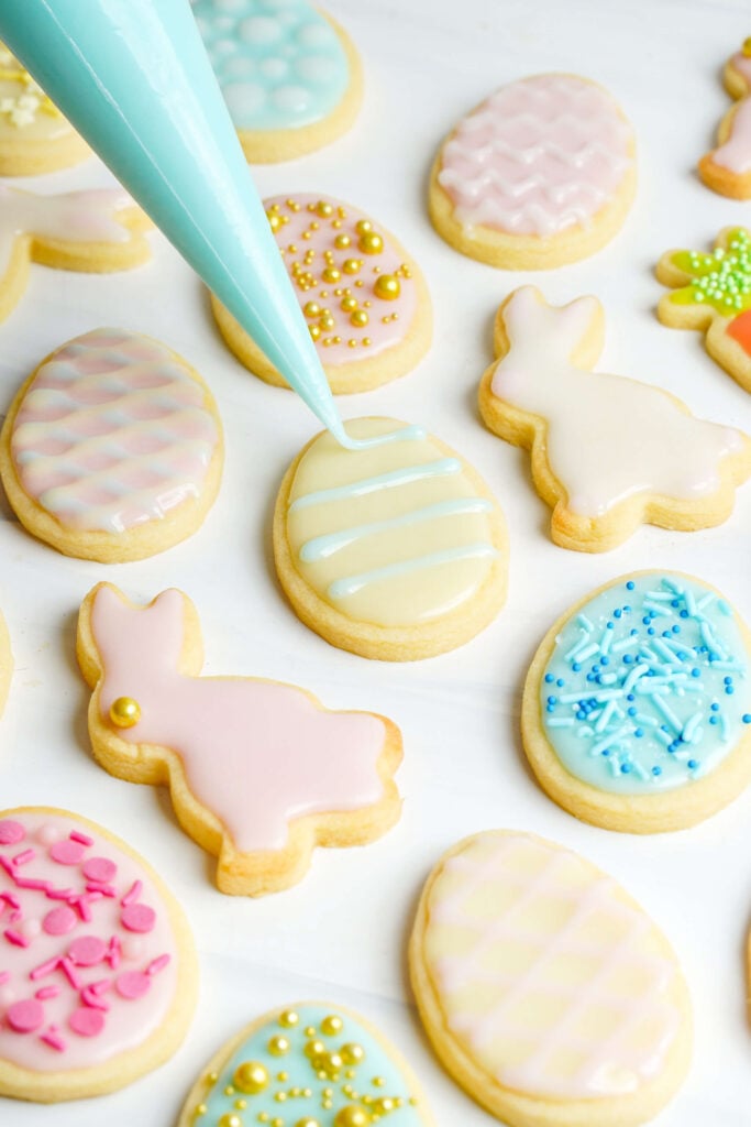 Glaçage bleu clair sur un biscuit en forme d'œuf de Pâques décoré entouré de biscuits pastel en forme de lapin, d'œuf et de carotte.