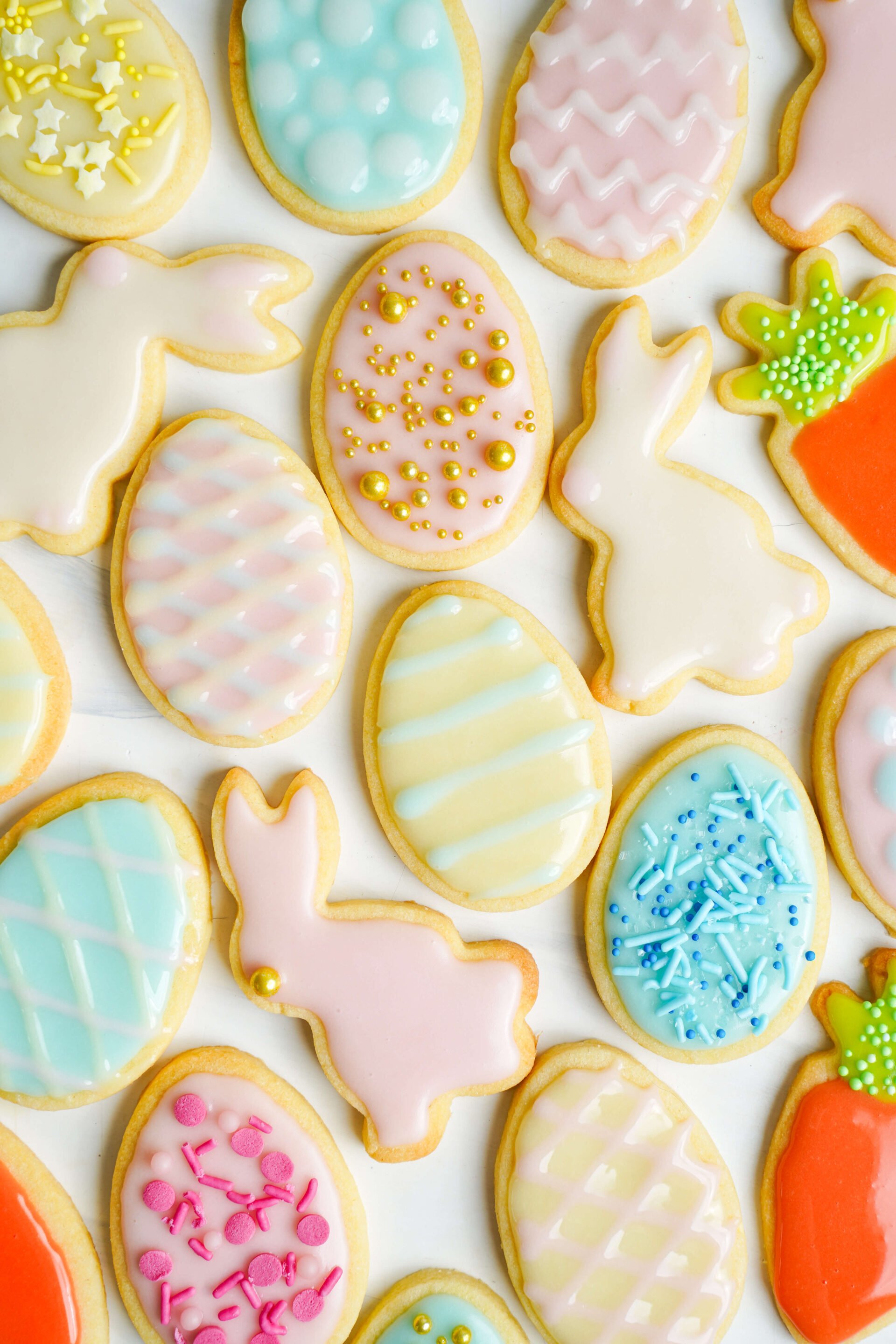 Un assortiment coloré de biscuits de Pâques décorés en forme d'œufs, de lapins et de carottes, disposés sur une surface blanche avec un glaçage pastel et des vermicelles.