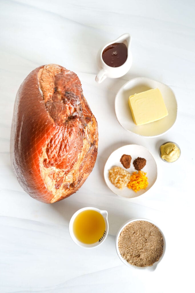 Vue d'ensemble des ingrédients pour une recette de jambon glacé au sucre brun, y compris le jambon avec os, du beurre, du sucre brun, du miel, de la moutarde de Dijon, de l'ail, des épices, du zeste d'orange et du bouillon de poulet