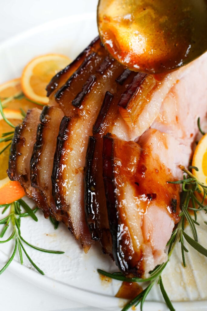 Jambon glacé tranché étant arrosé de glaçage sur une assiette blanche avec des tranches d'orange et du romarin