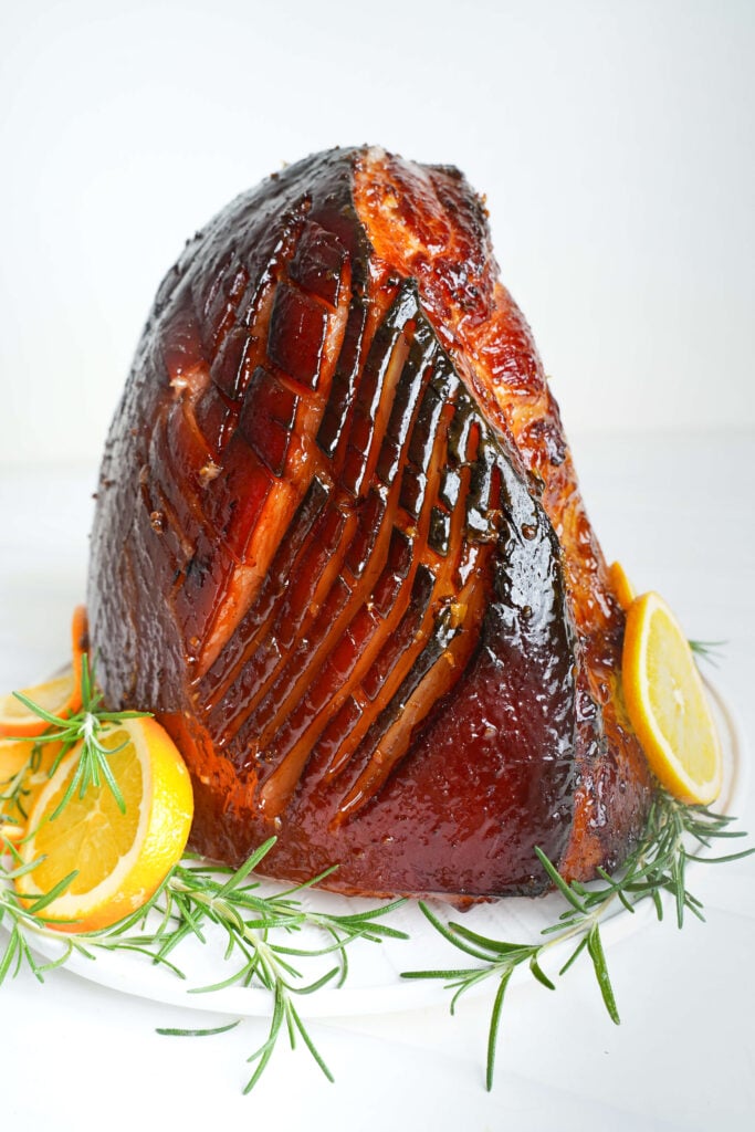 Jambon glacé au sucre brun entièrement cuit, tranché et servi avec des rondelles d'orange et des brins de romarin frais sur un plat blanc